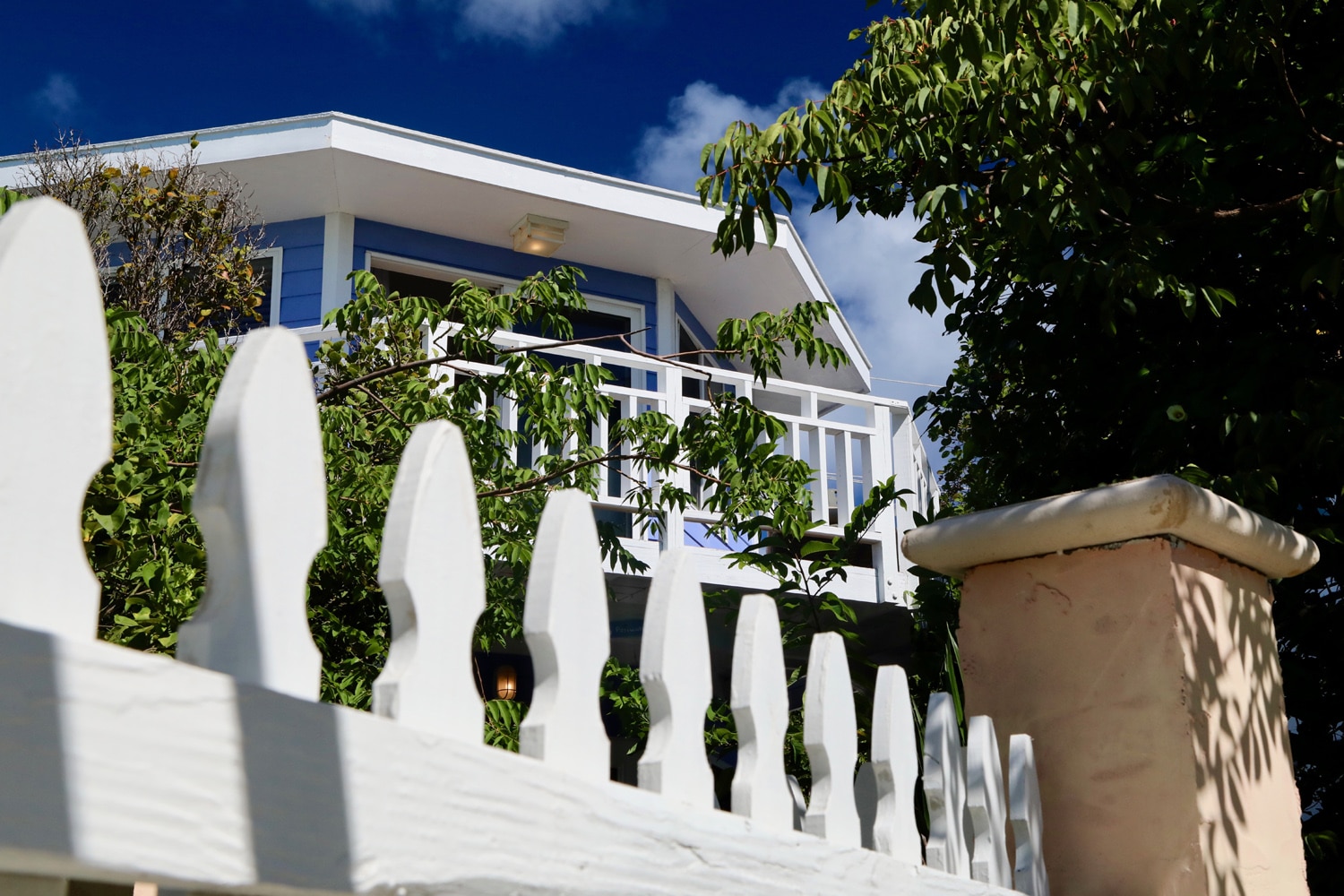 Staniel Cay Yacht Club A white picket fence frames the view of a house's balcony, surrounded by lush greenery under a clear blue sky, reminiscent of a serene escape at Staniel Cay Resort. Vacation Rentals and Resort on Staniel Cay Exuma