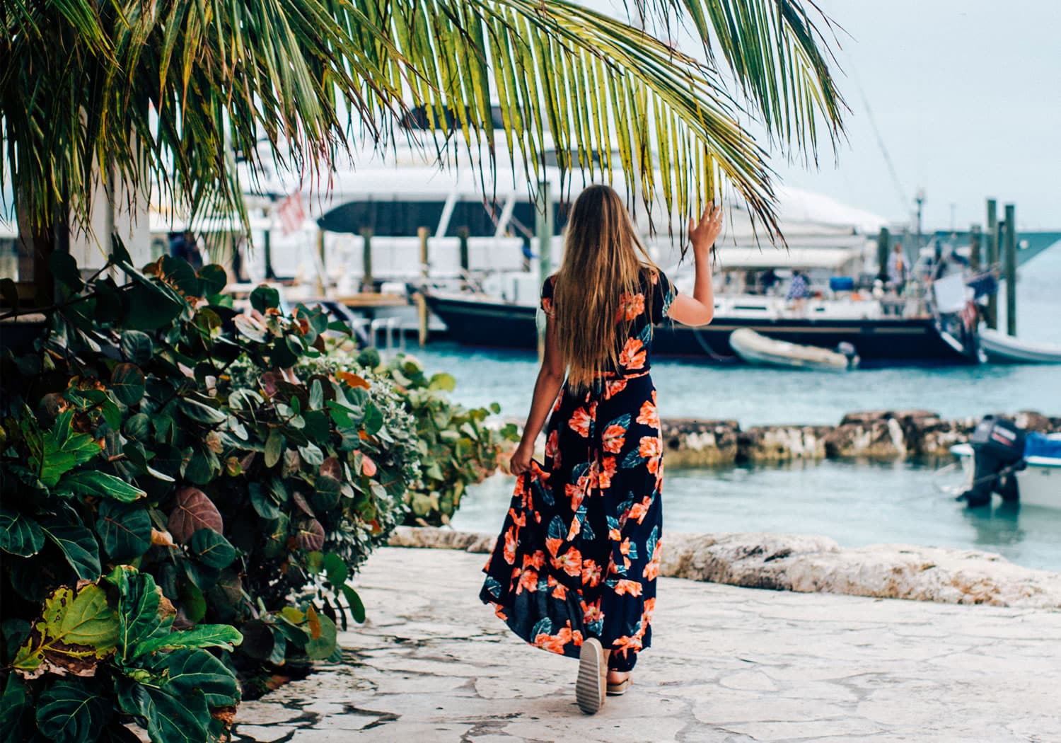 A woman in a floral dress walking down a path near a boat on her Staniel Cay vacation.