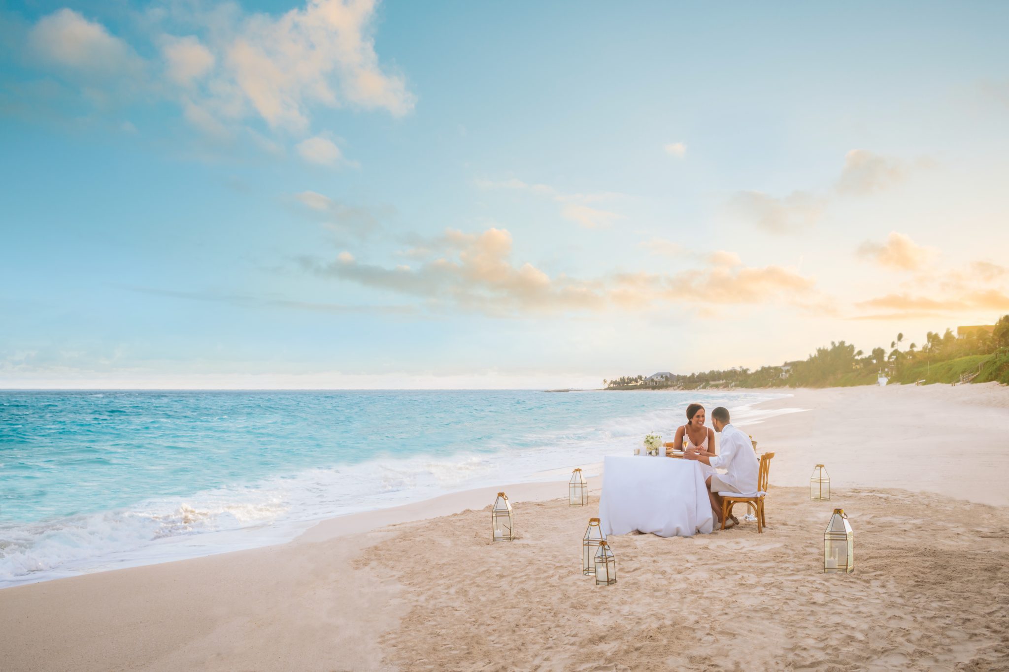 A couple enjoys a romantic evening at a table on the beach, surrounded by the mesmerizing sunset and the serene ambiance of Staniel Cay.