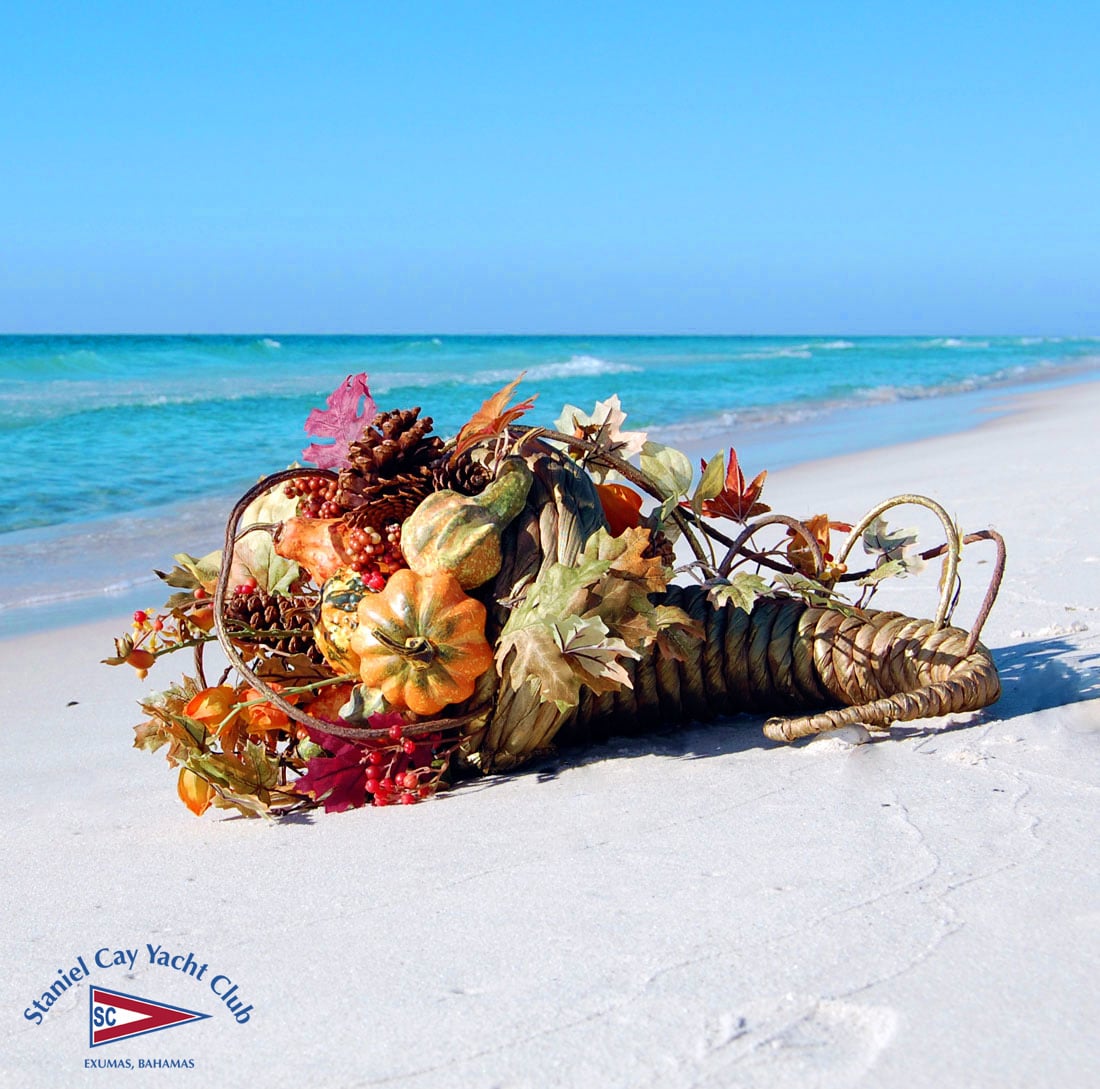Staniel Cay Yacht Club A cornucopia filled with autumn leaves, pumpkins, and pine cones rests on white sand by the ocean under a clear blue sky, celebrating Thanksgiving Dinner at Staniel Cay. The Staniel Cay Yacht Club logo appears in the corner. Vacation Rentals and Resort on Staniel Cay Exuma