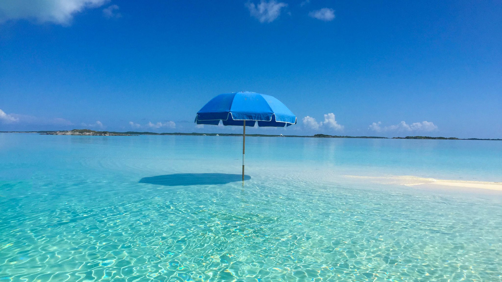 Staniel Cay Yacht Club A solitary blue beach umbrella stands in the shallow, clear turquoise waters of Exuma Cays under a bright blue sky. Vacation Rentals and Resort on Staniel Cay Exuma