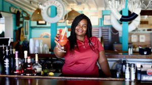 Staniel Cay Yacht Club Bartender in a red shirt holds up a pink cocktail with a straw and fruit garnish, standing behind a bar with various bottles and nautical-themed decor inspired by the Exuma Cays. Vacation Rentals and Resort on Staniel Cay Exuma