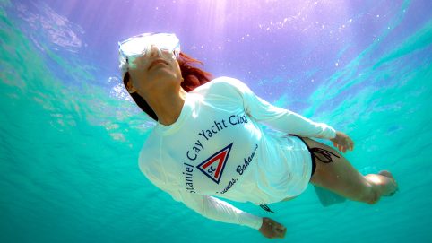 Staniel Cay Yacht Club A person snorkeling underwater in the clear blue waters of Exuma Cays, wearing a white shirt adorned with a yacht club logo and goggles. Sunlight dances through the surface above, creating a mesmerizing play of light. Vacation Rentals and Resort on Staniel Cay Exuma
