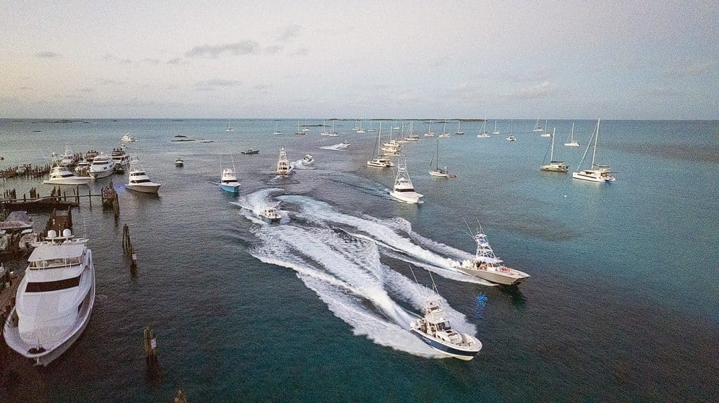 Staniel Cay Yacht Club Aerial view of motorboats speeding through turquoise water, leaving white trails, surrounded by multiple anchored yachts and sailboats during the annual SCYC Wahoo Blitz Fishing Tournament. Vacation Rentals and Resort on Staniel Cay Exuma