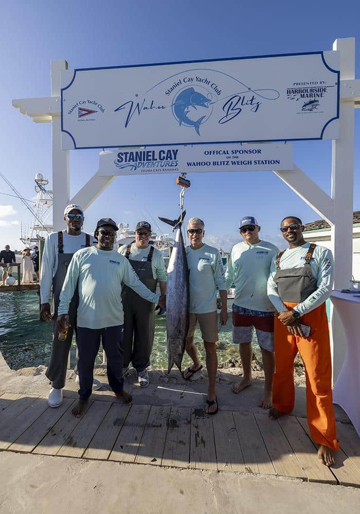 Staniel Cay Yacht Club Six men pose on a dock under a sign, holding a large fish at a weigh station during the SCYC Wahoo Tournament at Staniel Cay Yacht Club. Vacation Rentals and Resort on Staniel Cay Exuma