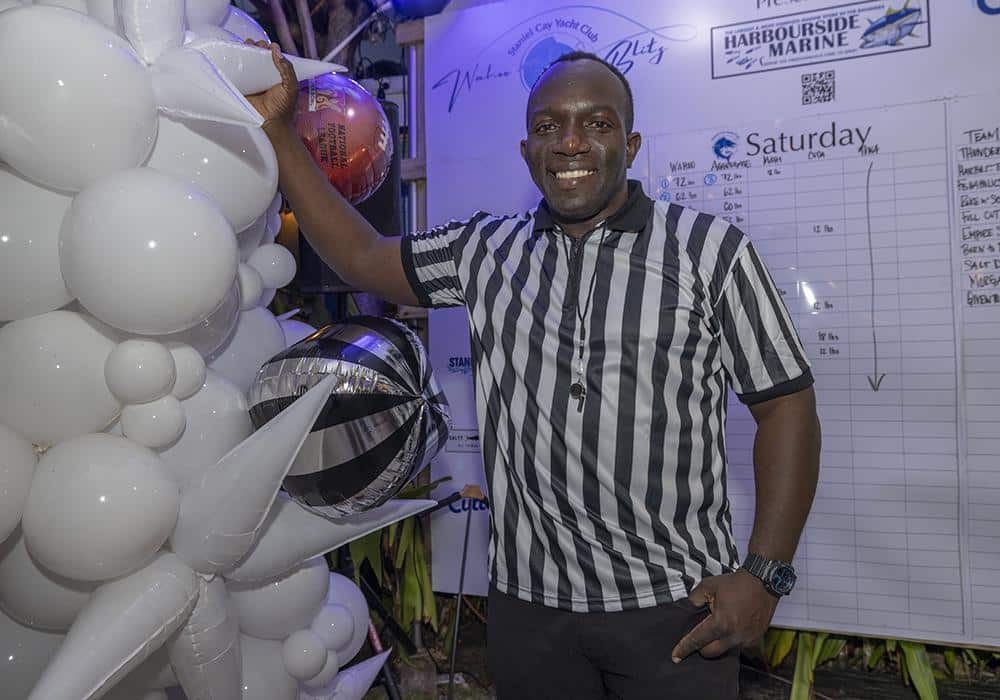 Staniel Cay Yacht Club Smiling man in a black and white referee shirt stands beside white balloons, holding a basketball, with a schedule board for the 2027 Wahoo Tournament at SCYC in the background. Vacation Rentals and Resort on Staniel Cay Exuma